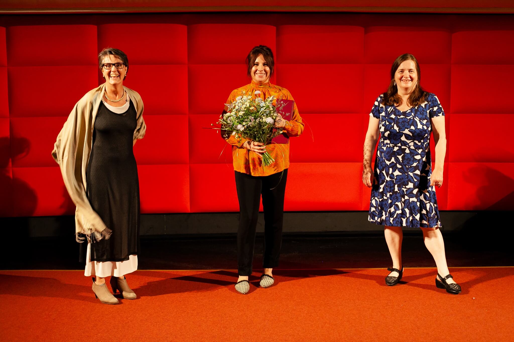 Drei Personen stehen vor einer roten Wand. Die Preisträgerin Alexandra Makarová in der Mitte hält einen Blumenstrauß.