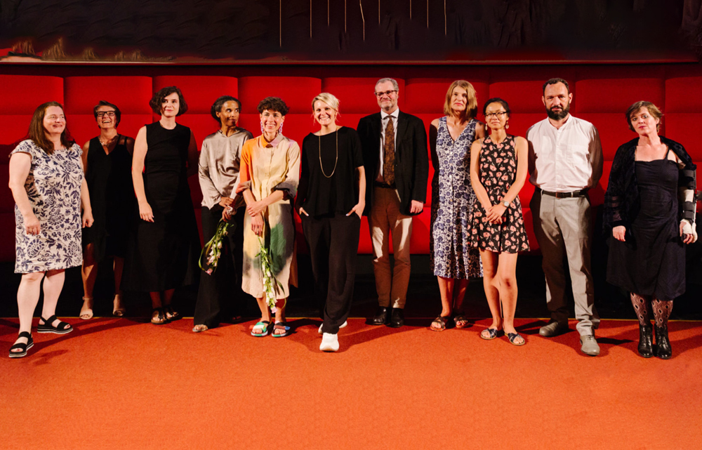 Ein Gruppenfoto von 11 Personen: Die Preisträger*innen und Jurymitglieder*innen vor einer roten Wand im Filmcasino.