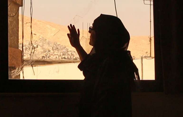 Filmstil aus "Chaos": Eine Frau mit Kopftuch in einem dunklen Raum legt ihre Hand auf die Fensterscheibe hinter welcher gelbe Wüstenlandschaft zu erkennen ist.