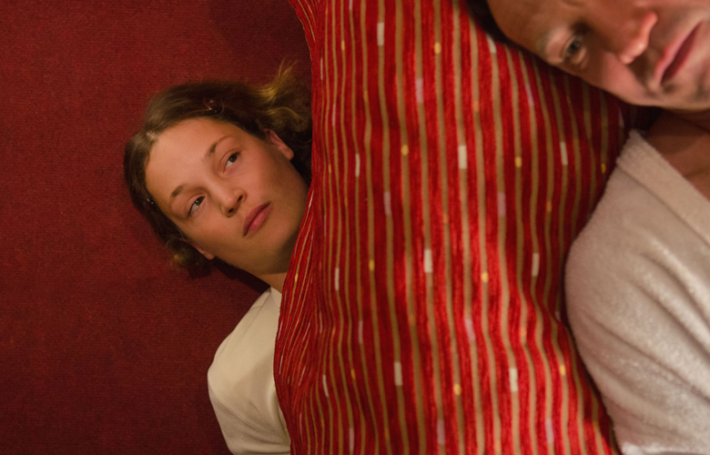 Filmstill aus "Das Zimmermädchen Lynn": Eine Vogelperspektive auf zwei liegende Personen, eine am roten Teppichboden und die andere etwas höher am roten Bettüberzug.