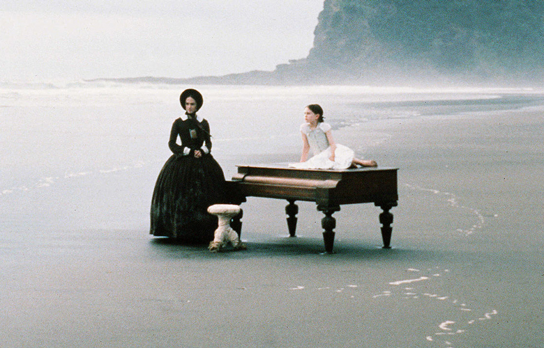 Filmstil aus "The Piano": Ein Klavier steht am Strand, ein Kind in weißem Kleid liegt am Klavier und eine Frau in schwarzem Kleid steht neben dem Klavier. Im Hintergrund ist das Meer und Klippen zu erkennen.